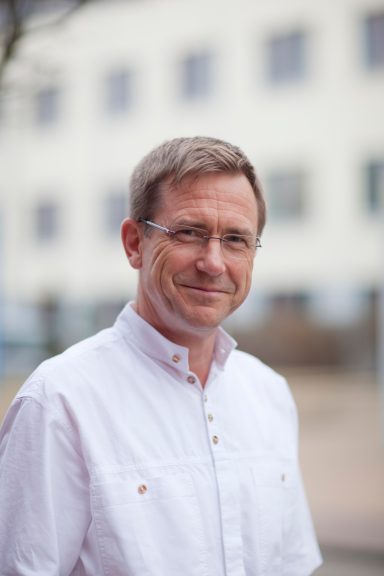 Lächelnder Mann in einem weißen Hemd vor einem unscharfen, hellen Hintergrund.