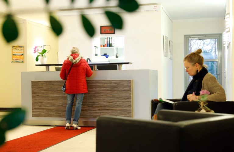 Eine Frau in rotem Mantel steht an einem Empfangstresen, während eine andere sitzt.