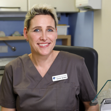 Lächelnde Frau in medizinischer Kleidung sitzt an einem Schreibtisch in einer Praxis.