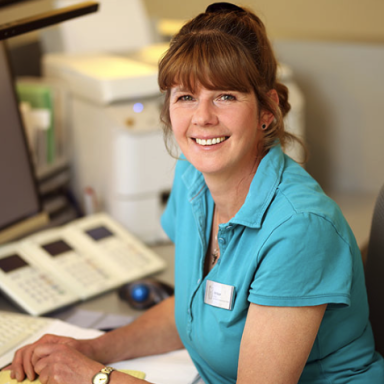 Frau mit dunklen Haaren, die an einem Schreibtisch sitzt und lächelt.